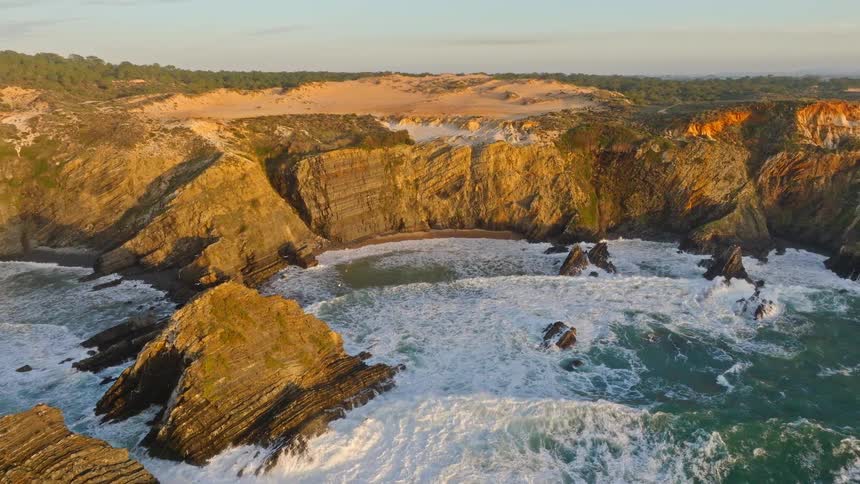 Stunning aerial pullback reveals a rugged coastline with dramatic cliffs, a hidden beach, and crashing waves at golden hour