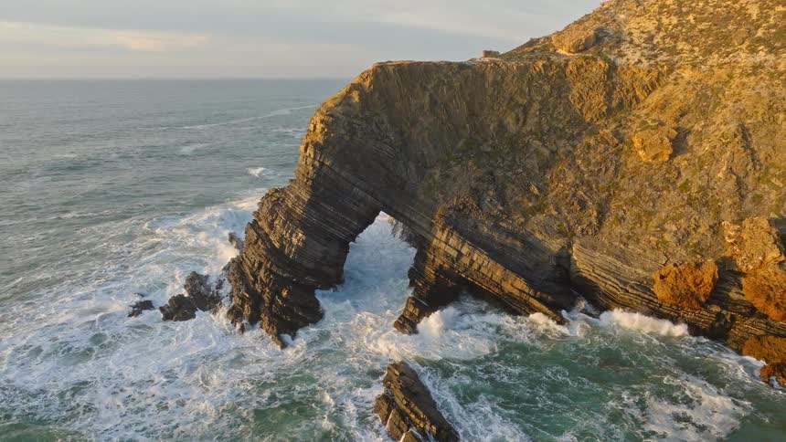 Dramatic aerial pullback reveals a massive natural rock arch battered by powerful ocean waves during a golden sunset