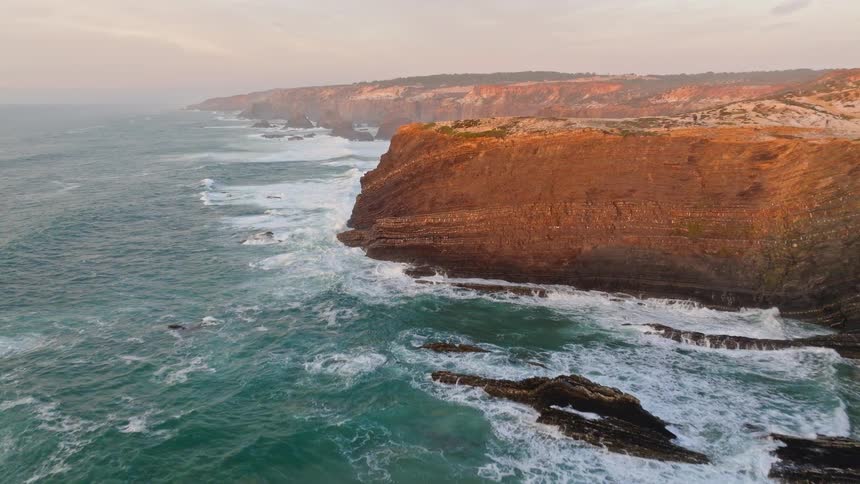 An aerial drone flies slowly over a dramatic and rugged coastline where powerful ocean waves crash against the rocky cliffs during a beautiful sunset
