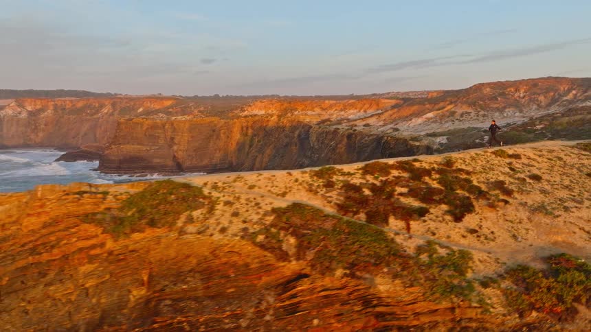 A lone man jogs along a scenic coastal trail on a cliffside, with an aerial pullback revealing the vast, epic landscape at sunset