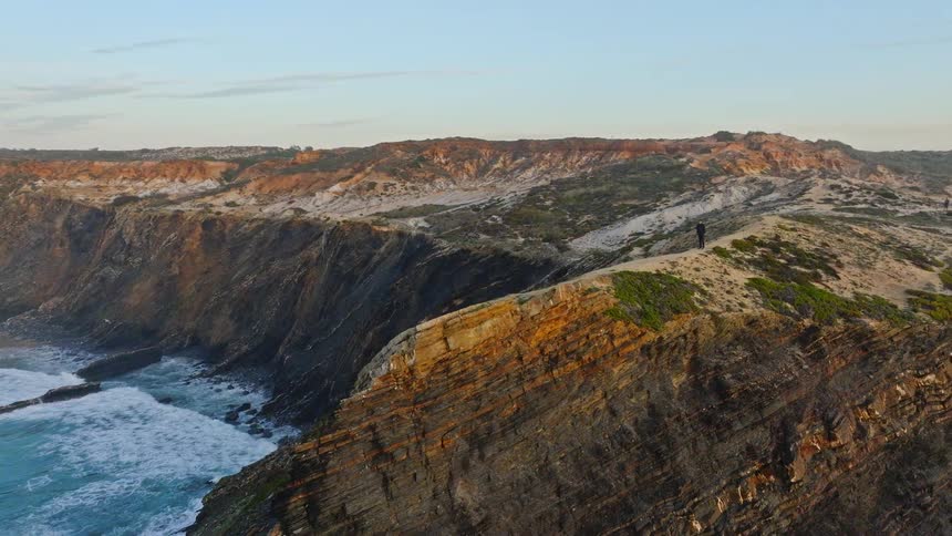 Aerial footage follows a lone runner along a spectacular coastal cliff, revealing a dramatic landscape with waves crashing against the shore