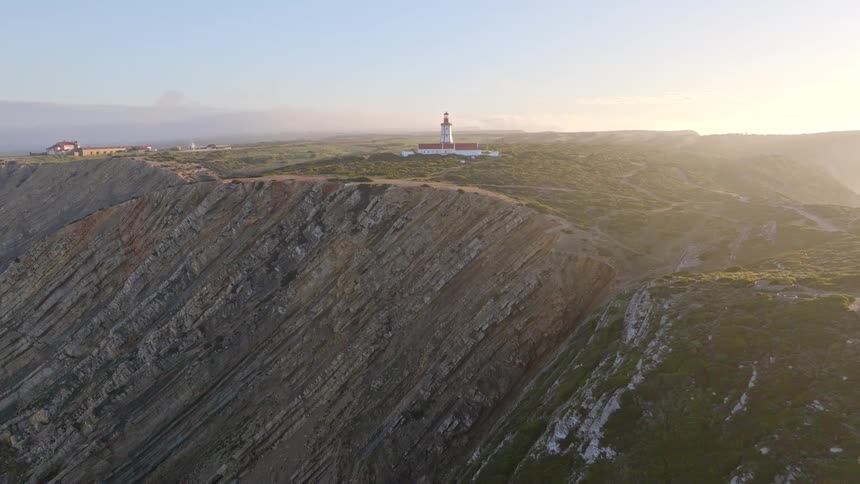 An aerial shot pulls back from a coastal lighthouse to reveal its stunning location on a dramatic, rocky cliff overlooking the ocean at sunrise