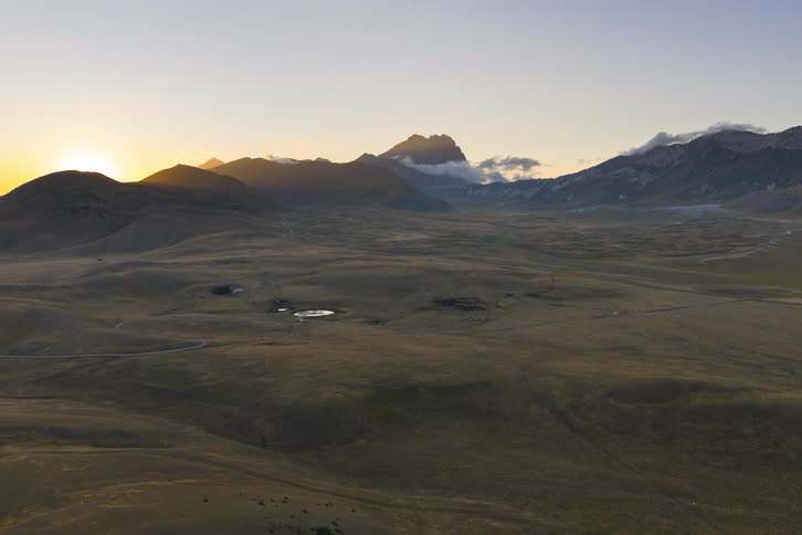 Golden hour aerial footage flies over rolling hills towards a vast mountain valley as the sun sets on the horizon