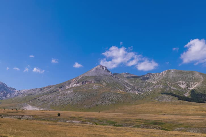 A timelapse of white clouds moving across a bright blue sky over a vast mountain range and a golden valley