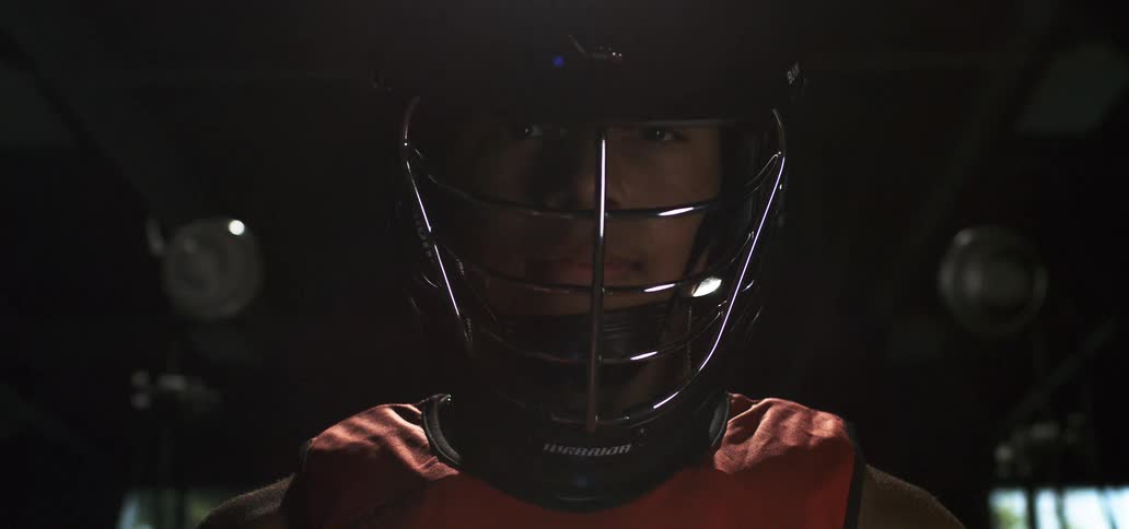 A dramatic close-up of a lacrosse player in a helmet, staring intently at the camera in a dark room as bright lights flare