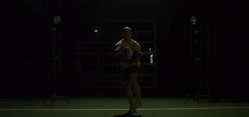 A male rugby player throws a spiral pass in super slow motion with dramatic lighting in a dark indoor facility