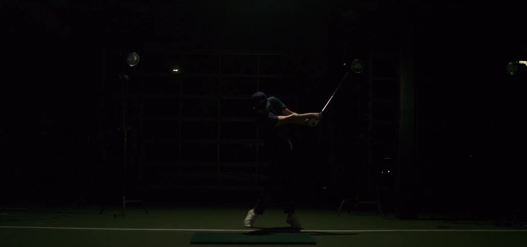 A male golfer's powerful swing is captured in dramatic slow motion as a bright light flashes in a dark indoor training facility