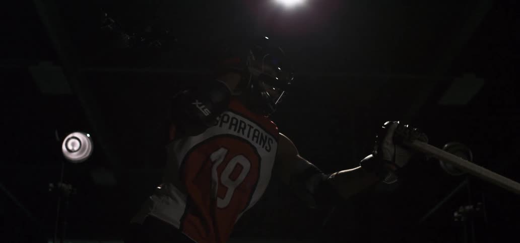 A lacrosse player stands dramatically in a dark room with spotlights as the camera slowly pans in super slow motion