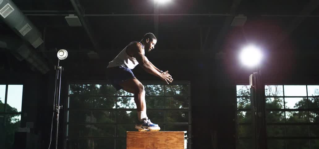 An athlete performs a powerful box jump in super slow motion, silhouetted by dramatic lighting in a gritty gym