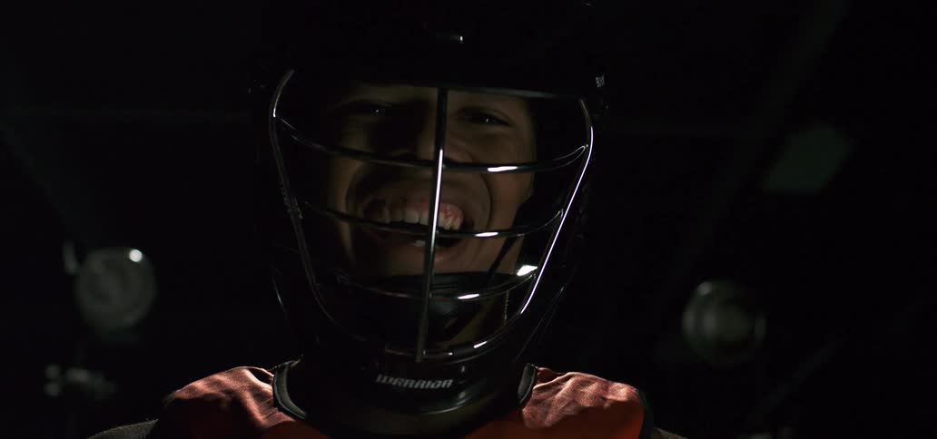 An intense lacrosse player wearing a helmet and pads smiles menacingly into the camera under a dramatic spotlight