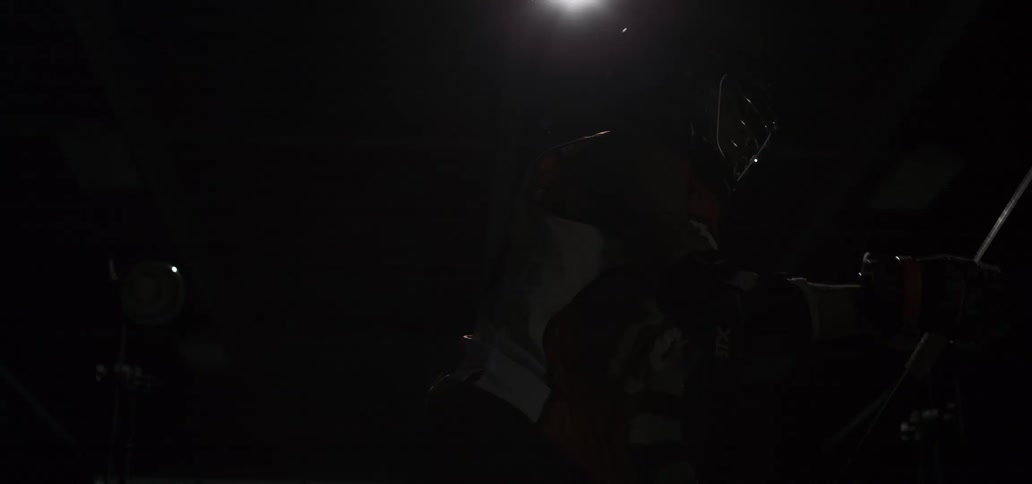 A lacrosse player in full uniform and helmet is dramatically revealed in a slow-motion studio shot as he holds his stick in a ready stance