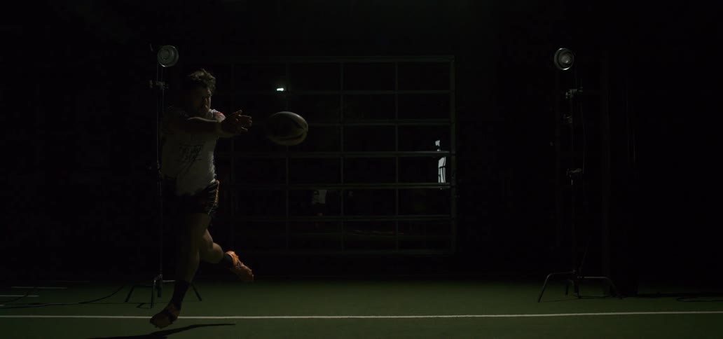 Rugby player throws a powerful spiral pass in dramatic slow motion, illuminated by a flash of bright studio lights