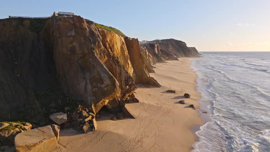 Golden hour light illuminates the massive cliffs and sandy shores of an expansive beach as gentle waves roll in