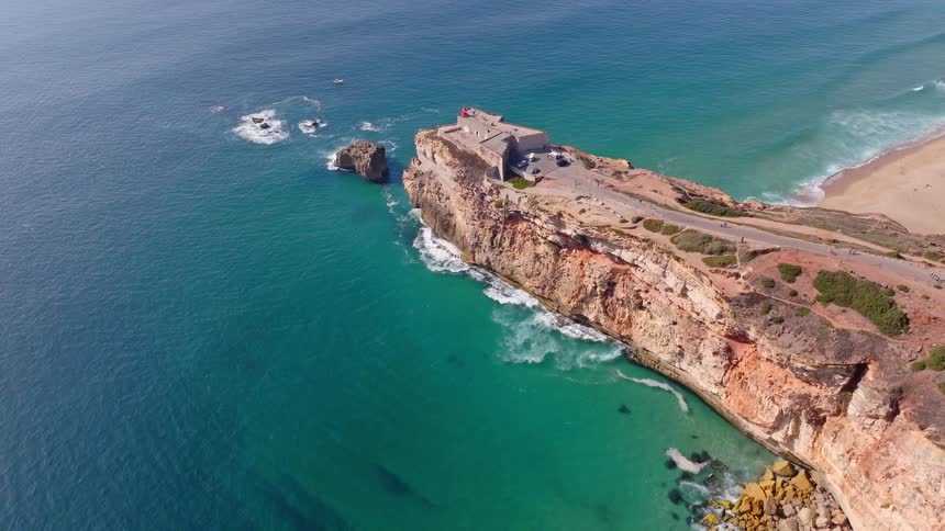 A majestic aerial pullback of a historic fort on a rugged cliff overlooking the stunning turquoise ocean in Nazaré, Portugal