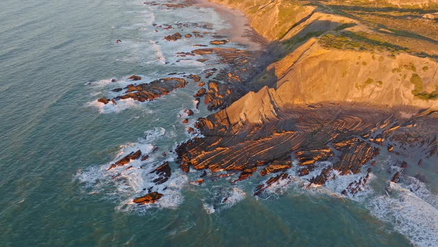 A serene aerial view of waves crashing against a rugged, layered rock coastline bathed in the warm glow of sunset