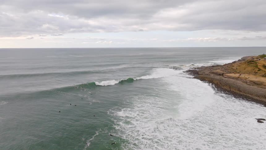 An aerial view shows surfers enjoying the ocean waves rolling towards a rugged coastline on a cloudy day
