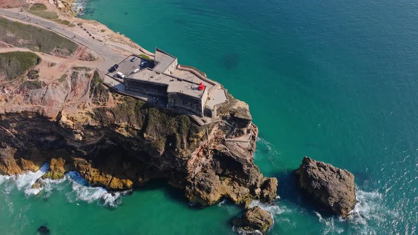 An aerial camera tilts up to reveal the stunning Fort of São Miguel Arcanjo lighthouse on a cliff overlooking the turquoise Atlantic Ocean