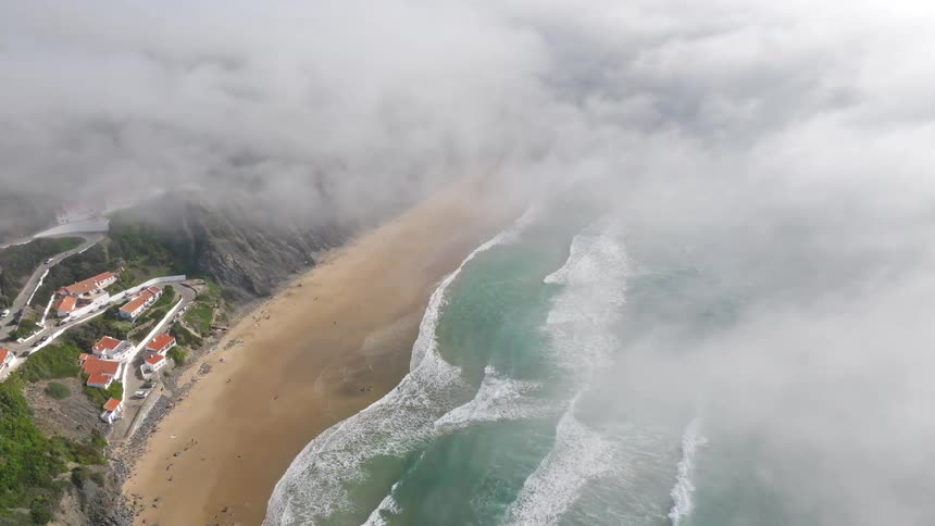 A majestic aerial shot rises through a sea of clouds, unveiling a hidden coastal town and a beautiful sandy beach with turquoise waves