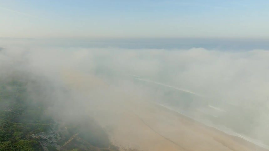 An ethereal aerial view flies through thick fog revealing camper vans parked on a cliffside above a vast sandy beach and ocean