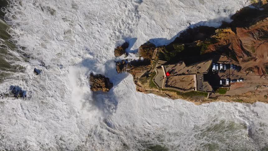 Stunning top-down aerial cinemagraph shows powerful, foamy ocean waves crashing against a rocky cliff with a fort where people are watching the spectacle