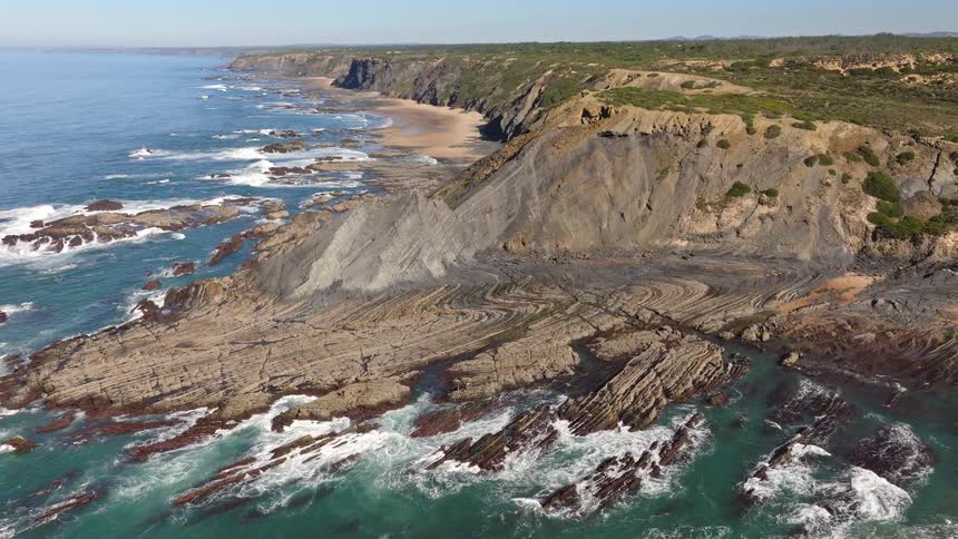 An incredible aerial view pulls back to reveal the stunning folded rock formations and rugged coastline of Zambujeira do Mar, Portugal