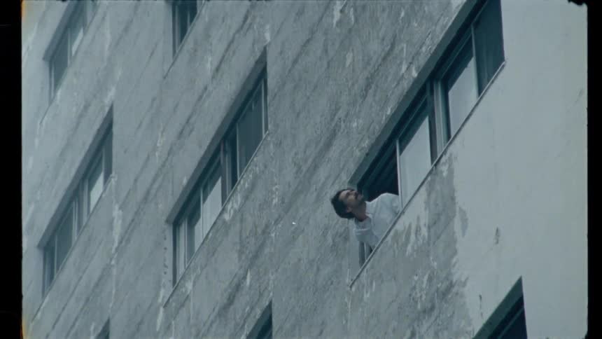 A man looks out the window of a stark, urban apartment building in a grainy, vintage-style shot that ends with an overexposed flash