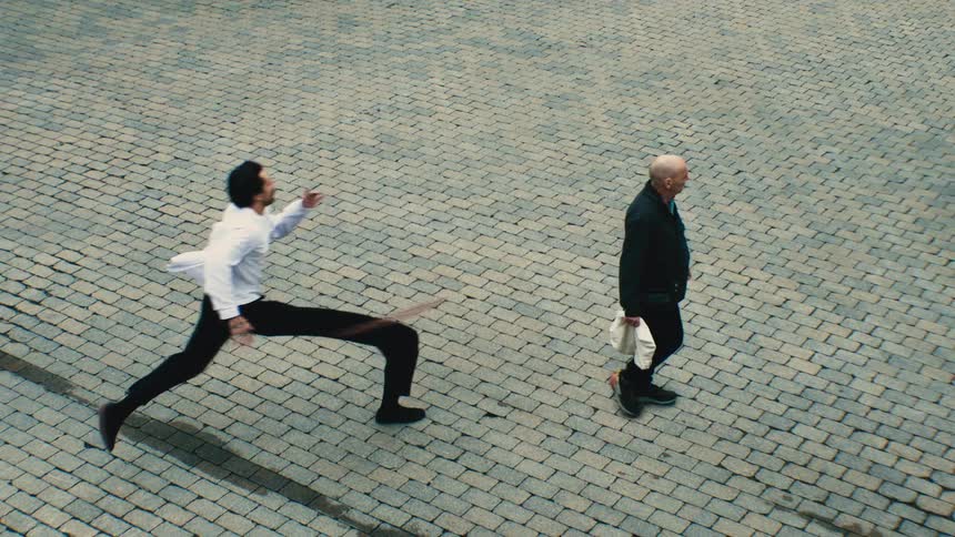 An artistic high-angle view shows a man's body and shadow distorting surrealistically as he runs in slow motion past a calmly walking older man on a cobblestone street