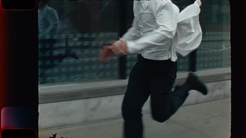 A determined man runs in slow motion down a city sidewalk, his reflection tracking alongside him in a vintage-style film shot