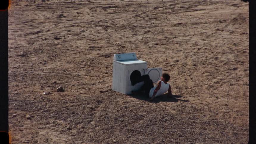A surreal vintage film-style clip shows a man emerging from a washing machine in the middle of a desolate quarry