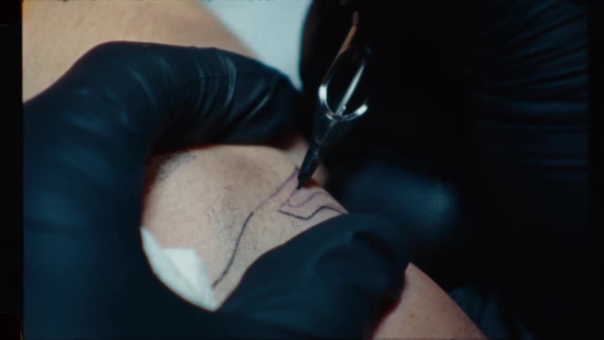 Vintage-style close-up shot shows a tattoo artist's gloved hands carefully inking a precise outline onto a client's skin