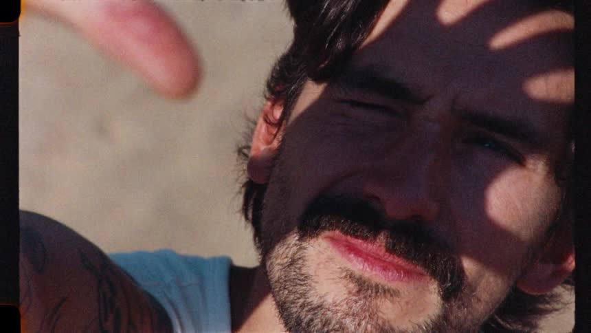 Vintage-style close-up footage shows a tattooed man with a mustache enjoying a sunny day, shielding his eyes from the sun