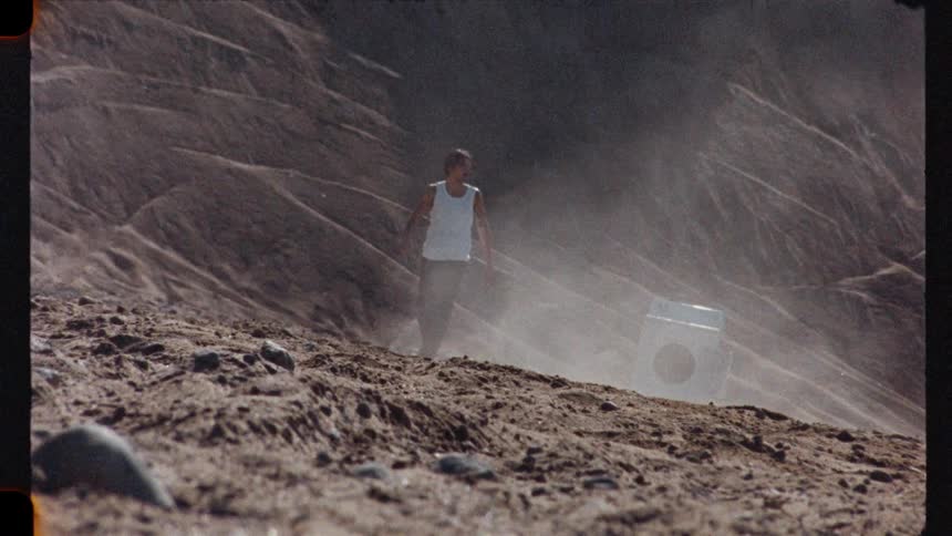 Vintage footage shows a man in a tank top standing next to a washing machine in a surreal, dusty, and windswept landscape