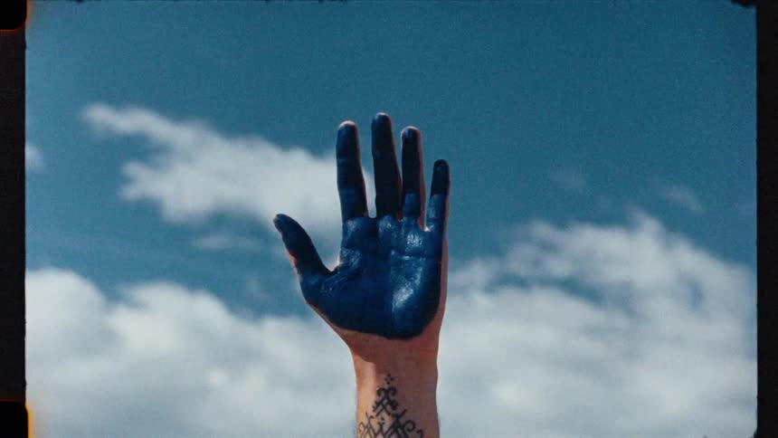 Close-up of a tattooed hand covered in blue paint opening towards the cloudy blue sky, shot on vintage film