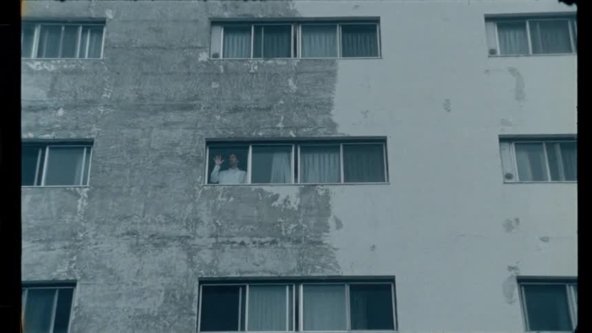 A man looks out the window of a drab, concrete apartment block, his solitary figure emphasized by a slow zoom out