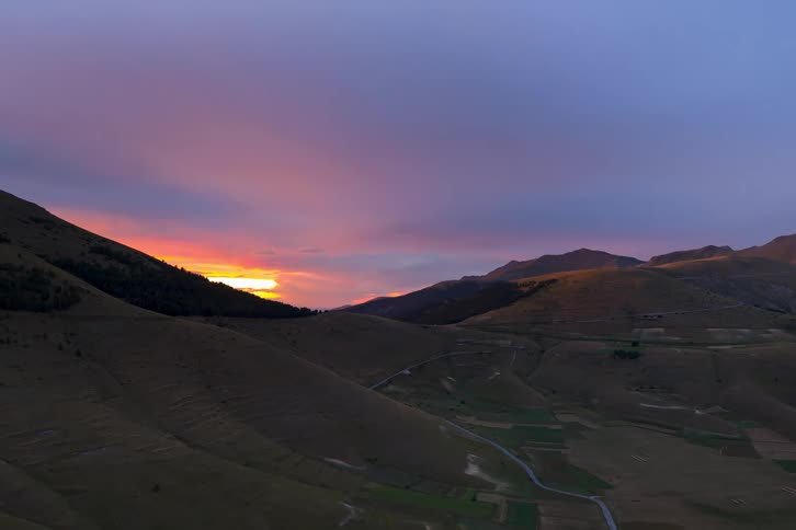 A stunning timelapse captures a vibrant pink and purple sunset over a vast mountain valley with rolling hills