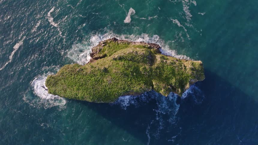 A mesmerizing top-down aerial view shows a lush green rocky islet surrounded by the gentle waves of the turquoise sea