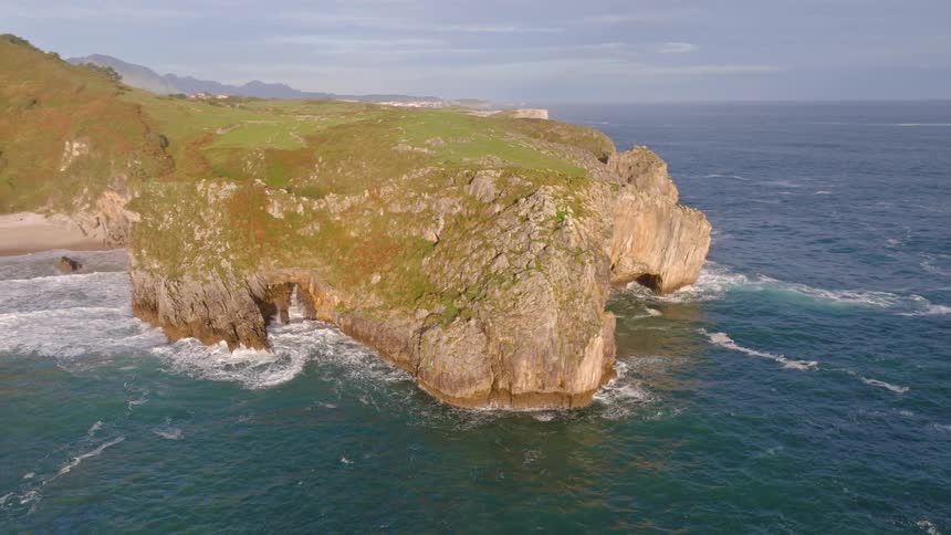 Majestic aerial shot pans across a rugged coastline, revealing dramatic green cliffs, natural rock arches, and powerful ocean waves below