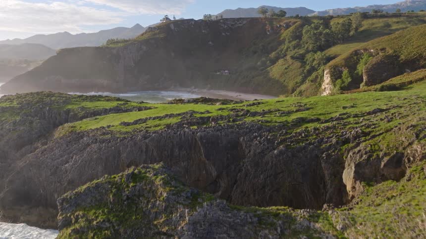 An epic aerial journey flies over a rugged, green coastline with a natural arch, revealing a secluded sandy beach in a hidden cove