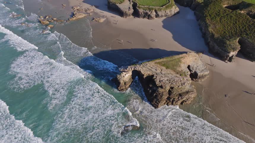 An aerial top-down shot pulls back to reveal the stunning natural rock arches and dramatic cliffs of a beautiful coastline as turquoise waves crash on the sandy beach