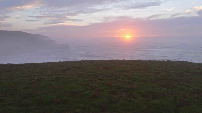 An aerial view reveals a couple watching a beautiful, misty sunset over the ocean from the roof of their camper van parked on a remote cliffside