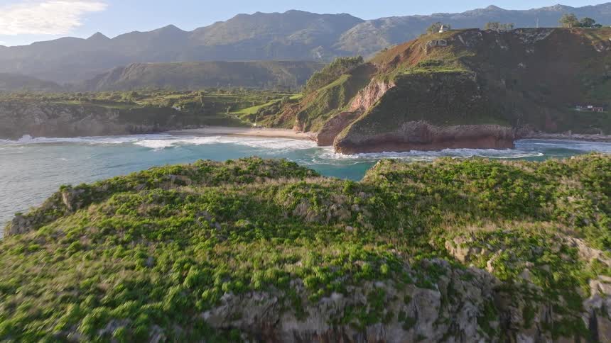 An aerial drone shot flies over a majestic green-topped rocky cliff to reveal a stunning hidden cove and sandy beach along the coast