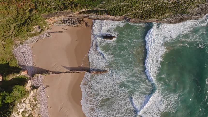 A static aerial top-down view shows powerful turquoise ocean waves crashing onto a secluded sandy beach within a rocky cove