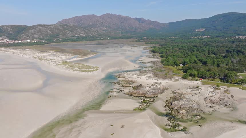 Stunning aerial view slowly ascending over a vast sandy beach at low tide, with a dense green forest and a mountain range in the background