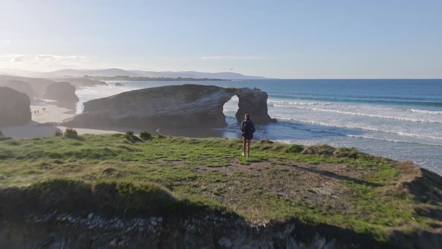 Aerial shot follows a female hiker to the edge of a cliff, revealing the stunning natural rock arches of As Catedrais beach at sunrise