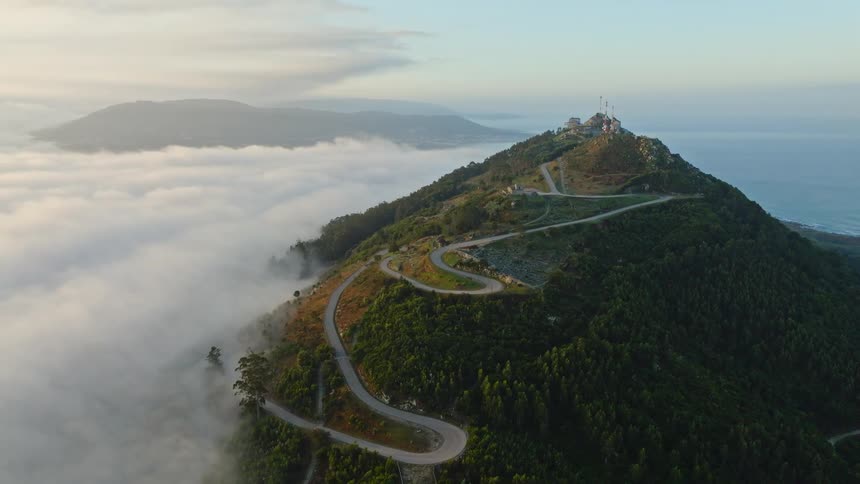An aerial view captures a winding road on a coastal mountain peak rising majestically above a sea of clouds