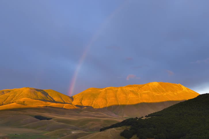 A colorful rainbow arches over sun-kissed golden mountains under a dramatic, cloudy sky as time passes
