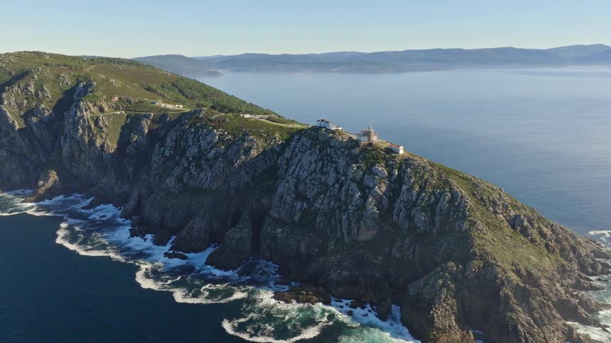 A scenic aerial pull-back shot reveals the majestic Cape Finisterre lighthouse situated on a rugged, rocky cliffside overlooking the ocean