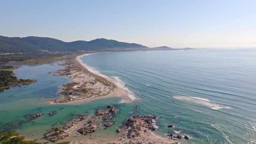 A stunning aerial pullback reveals a vast coastal landscape with a sandy spit separating a turquoise lagoon from the open ocean