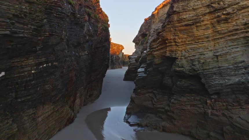 An awe-inspiring aerial flight through a narrow rock canyon on a sandy beach, emerging to reveal a stunning coastline at golden hour