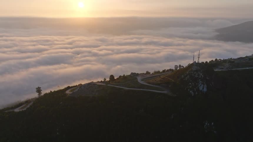 An incredible aerial view flies over a mountain peak, revealing a winding road and a breathtaking sea of clouds during a golden sunrise
